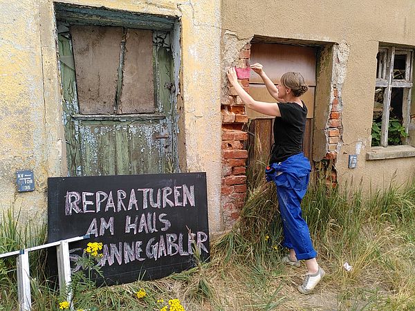 Susanne Gabler steht steht an einem verfallenen Haus. Davor steht ein Schild mit der Aufschrift "Reparaturen am Haus Susanne Gabler".