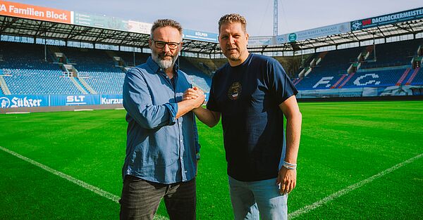 Zwei Männer stehen im Stadion auf dem Rasen und reichen sich die Hände.