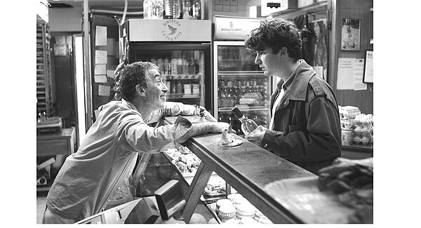 Eine Ladentheke. Davor steht ein junger Mann mit einer Weinflasche in der Hand. Dahinter ein älterer Mann. Beide schauen sich an.