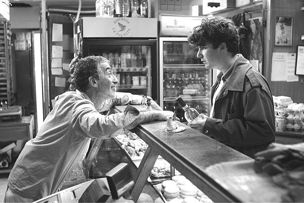 Eine Ladentheke. Davor steht ein junger Mann mit einer Weinflasche in der Hand. Dahinter ein älterer Mann. Beide schauen sich an.