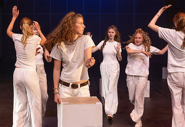 Sechs Mädchen stehen auf einer Bühne und tanzen. Alle tragen weiße Kleidung.