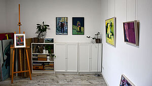 Das Foto zeigt einen Raum mit Malereien, einer Staffelei und Malzeug. 