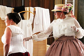 Eine Frau in historischer Kleidung zieht einer anderen Frau das Korsett fest.