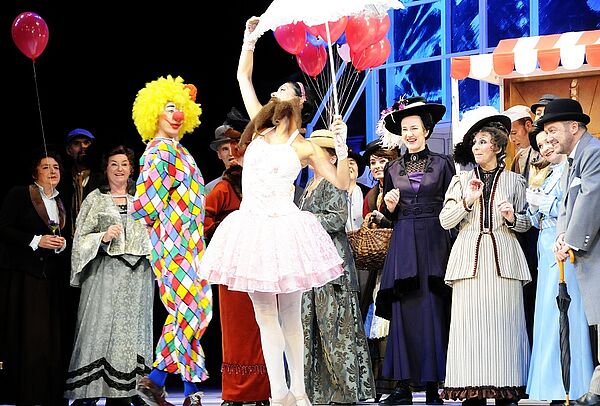 15 Schauspielerinnen und Schauspieler stehen in bunten Kostümen auf einer Bühne. Mit dabei: ein Clown, eine Ballerina, feine Ladys und vornehme Herren.