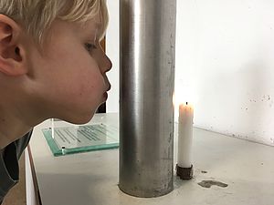 Ein Junge pustet auf ein Metallrohr. Dahinter steht eine brennende Kerze.