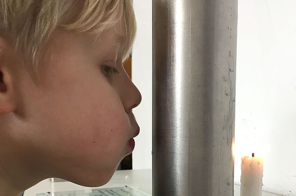 Ein Junge pustet auf ein Metallrohr. Dahinter steht eine brennende Kerze.