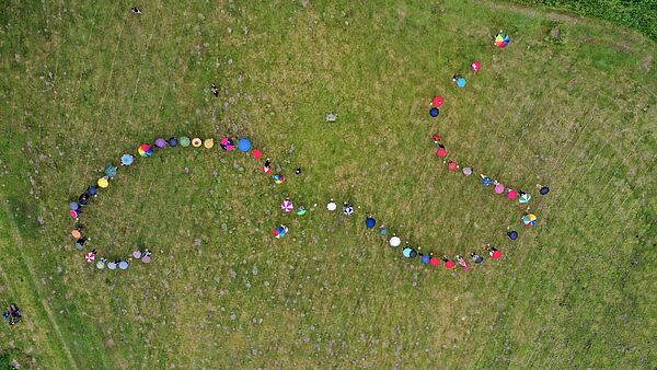 Luftaufnahme von einer Wiese. Darauf stehen mehr als 60 Menschen mit aufgespannten Regenschirmen in einer Schlangenlinie. Von oben sieht man nur die bunten Schirme.