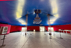 Ein großer Raum. Die Wände sind blau und rot. Ein paar Tische stehen schon. An den Zeltwänden stehen Bänke.