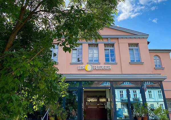 Ein zweistöckiges Gebäude. Im Erdgeschoss befindet sich ein Glaseingang. An der Fassade steht Luna Filmtheater. Links ragen Blätter eines Baumes ins Bild.