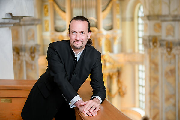 Ein Mann in schwarzem Anzug schaut in die Kamera. Im Hintergrund eine Kirchensaal. 