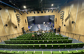 Grüne Stühle bilden absteigend neun Sitzreihen. Jede Reihe hat zwischen 14 und 16 Plätze. Die Wände sind holzvertäfelt. An ihnen hängen Lautsprecher und Scheinwerfer. Die Bühne ist von Holz umrahmt. Auf der Bühne stehen Mitarbeiter des Theaters, Fotografen und Journalisten.