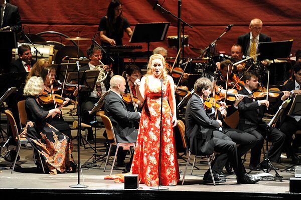 Eine Solistin steht in einem roten, langen Kleid an einem Mikrofon und singt. Hinter ihr spielt das Orchester.