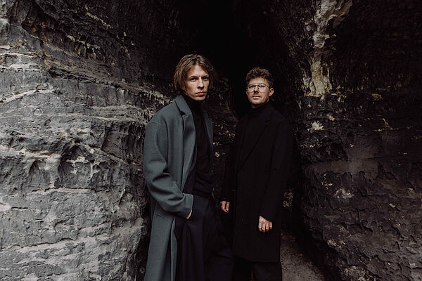 Zwei junge Männer stehen in Mänteln an einer Wand und gucken cool in die Kamera.