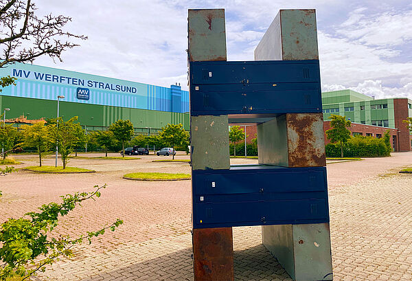 Im Vordergrund steht das Kunstwerk. Der Künstler hat abwechseln Spinde und Stahlblöcke übereinander gestapelt, in der Mitte fehlen die Blöcke. Dadurch entstehen Freiräume zum Durchgucken. Hinter dem Kunstwerk stehen Werftgebäude.