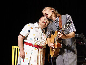 Eine Frau und ein Mann stehen auf einer Bühne. Sie lehnen ihre Köpfe aneinander. Der Mann spielt Gitarre.