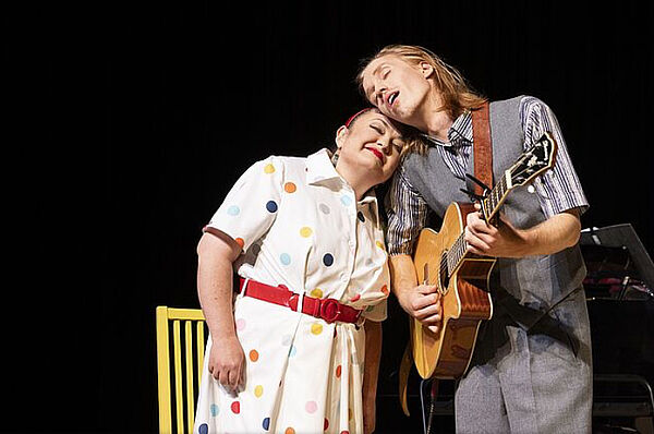 Eine Frau und ein Mann stehen auf einer Bühne. Sie lehnen ihre Köpfe aneinander. Der Mann spielt Gitarre.