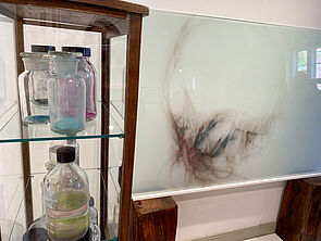 In einer Vitrine stehen Gläser und Flaschen. Daneben hängt ein Glasbild.