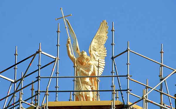 Der Erzengel ist eine goldene Figur. Sie steht umgeben von blauem Himmel. Links und rechts von ihr stehen Gerüste.