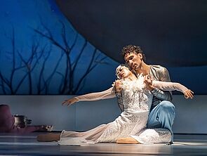 Ein Mann und eine Frau. Sie tragen Kostüme. Sie knien eng aneinander auf einer Bühne. 