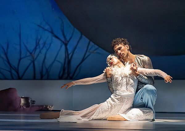 Ein Mann und eine Frau. Sie tragen Kostüme. Sie knien eng aneinander auf einer Bühne. 