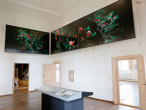 Ein Ausstellungsraum mit drei geöffneten Türen. In der Mitte steht eine Vitrine. Über den Türen hängen drei großformatige dunkle Fotografien an der Wand. Sie zeigen rote Beeren an einem Strauch. Der Hintergrund ist schwarz.