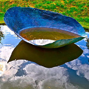 Eine Skulptur. Sie erinnert an eine Schale oder ein nach oben gebogenes Blatt und ist mit Wasser gefüllt.