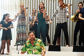 Schauspieler Martin Neuhaus steht im Plenarsaal am Rednerpult. Hinter ihm spielen Musiker Geige und Saxophon.