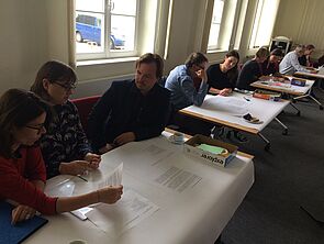 Neun Frauen und Männer sitzen an Tischen und lesen die Zettel, die vor ihnen liegen.