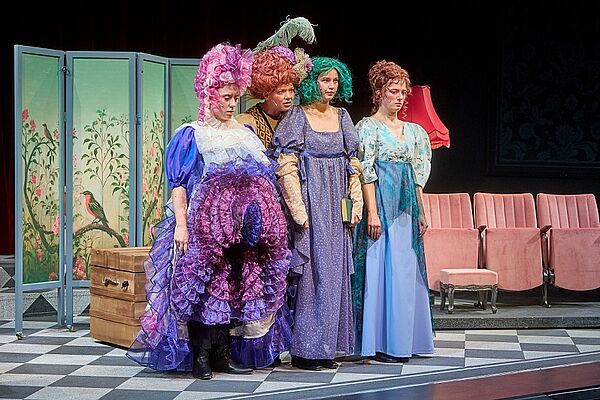 Vier Frauen mit Perücken und bunten Kleidern sehen auf der Bühne.