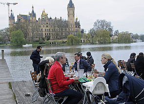Auf einer Terrasse am Schweriner See sitzen Frauen und Männer an Tischen. Im Hintergrund steht das Schweriner Schloss.