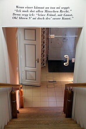 Eine Treppe führt nach unten. Über der Treppe steht: "Wenn einer kümmt un tau mi seggt: "Ick mak dat allen Minschen Recht." Denn segg ick: "leiwe Fründ, mit Gunst, Oh! lihren S' mi doch des' swore Kunst."