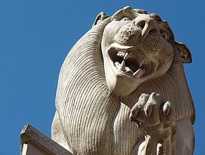 Ein Löwe aus Stein schaut von einem Sockel herab. Das Maul ist geöffnet, die Schneidezähne sind deutlich sichtbar. Seine linke Pfote ist erhoben. Um ihn herum: blauer Himmel.