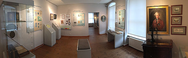 Ein Ausstellungsraum. An den Wänden hängen Gemälde und Infotafeln. Im Raum stehen zwei Hörstationen und eine Glasvitrine mit Ausstellungsstücken.