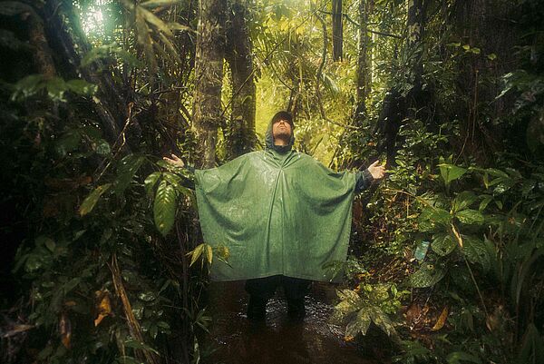 Marteria trägt einen grünen Regenponcho. Er steht in einem Wald, breitet die Arme aus und schaut nach oben.