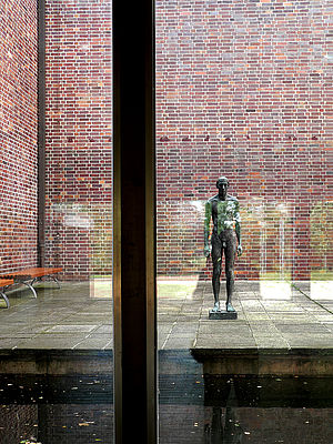 Skulptur eines nackten Mannes von vorn, fotografiert durch eine Fensterscheibe.