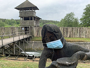 Eine Holzfigur trägt eine Corona-Schutzmaske im Gesicht.