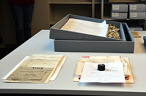 Auf einem hellgrauen Tisch liegt eine geöffnete Archivbox mit säurefreiem Verpackungsmaterial. Davor zwei Lagen historischer Dokumente, darunter ein altes Flurbuch mit der Aufschrift „Acten".