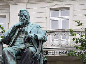 Fritz Reuter sitzt auf einem Stuhl. Eine Hand hat er ans Kinn geführt. Auf seinen Knien liegt ein Buch, das er mit einer Hand aufblättert. Das Denkmal steht auf einem Sockel. Dahinter befindet sich das Literaturmuseum.