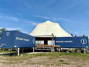 Hinter einem Zelt stehen au einem Gestell zwei große Container. Sie sind über eine Außenbühne miteinander verbunden. 