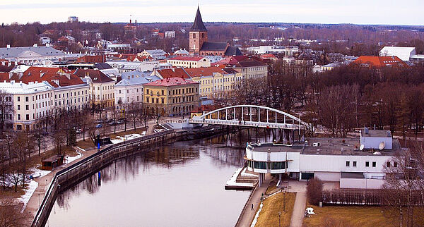 Ein Luftbild von Tartu. Im Vordergrund befindet sich ein Fluss mit einer Brücke. Dahinter stehen Häuser mit farbigen Fassaden. Zwischen ihnen ragt eine Kirche hervor. Am Horizont befindet sich Wald. 