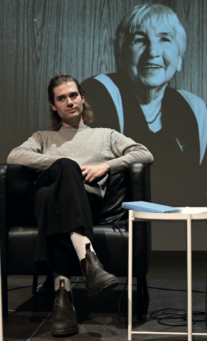 Ein Mann sitzt auf einem Stuhl. Im Hintergrund ist das Bild einer Frau zu sehen.