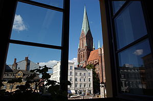 Der rechte Flügel eines Fensters im Rathaus ist geöffnet. Der Blick fällt auf den Marktplatz. Der Himmel ist blau. Zu sehen sind der Schweriner Dom, ein weißes historisches Gebäude und ein Fachwerkhaus.