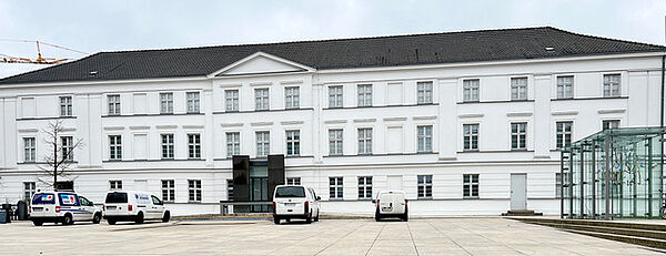 Ein Platz vor einem weißen, langgezogenen, dreistöckigen Haus.