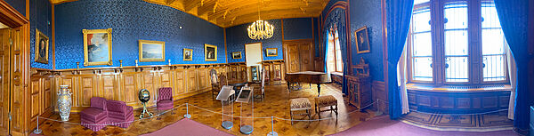 Ein Raum. Die Wände sind halb hoch mit Holz vertäfelt. Die anderen Stellen blau gestaltet. Im Raum stehen eine Bodenvase, pinkfarbene Sessel, ein Tisch mit Stühlen, Hocker und ein Klavier. An der Decke hängt ein Kronleuchter.