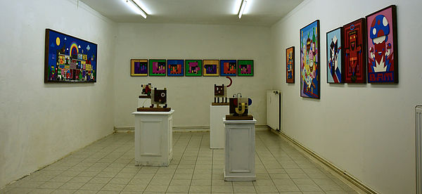 In einem hellen Raum stehen vier Sockel mit Skulpturen. An den Wänden hängen bunte, farbintensive Bilder.