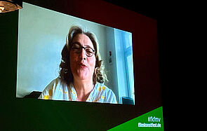 Eine Leinwand. Sie zeigt eine Frau. Die Frau spricht. 