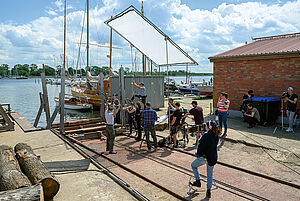 Ein Hafengelände. An der Hafenkante liegen Boote. An der Hafenkante stehen Mitarbeiter vom Filmteam mit Kameras und Mikrofonen. Sie filmen eine Szene. 