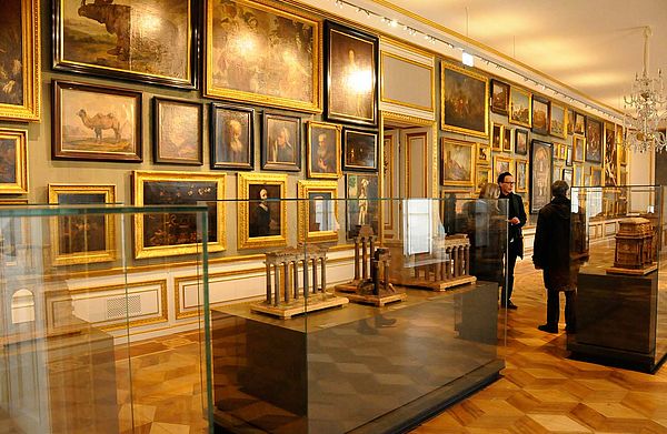 Ein Ausstellungsraum. Die Wand hängt voller Gemälde. Im Raum stehen Glasvitrinen, vor denen Menschen stehen.