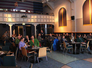 Ein Saal voller Tische und Stühle. An den Tischen sitzen Menschen. 