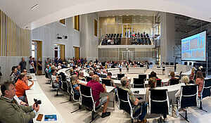 In einem großen Saal, dem Plenarsaal, steht eine große Leinwand. Darauf sind Bilder von Schwerin. Im Plenarsaal sitzen viele Menschen und schauen auf die Leinwand.
